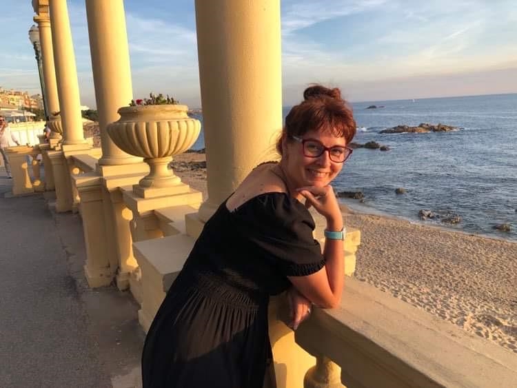 Monica is smiling at the camera, leaning over a balcony overlooking a beach