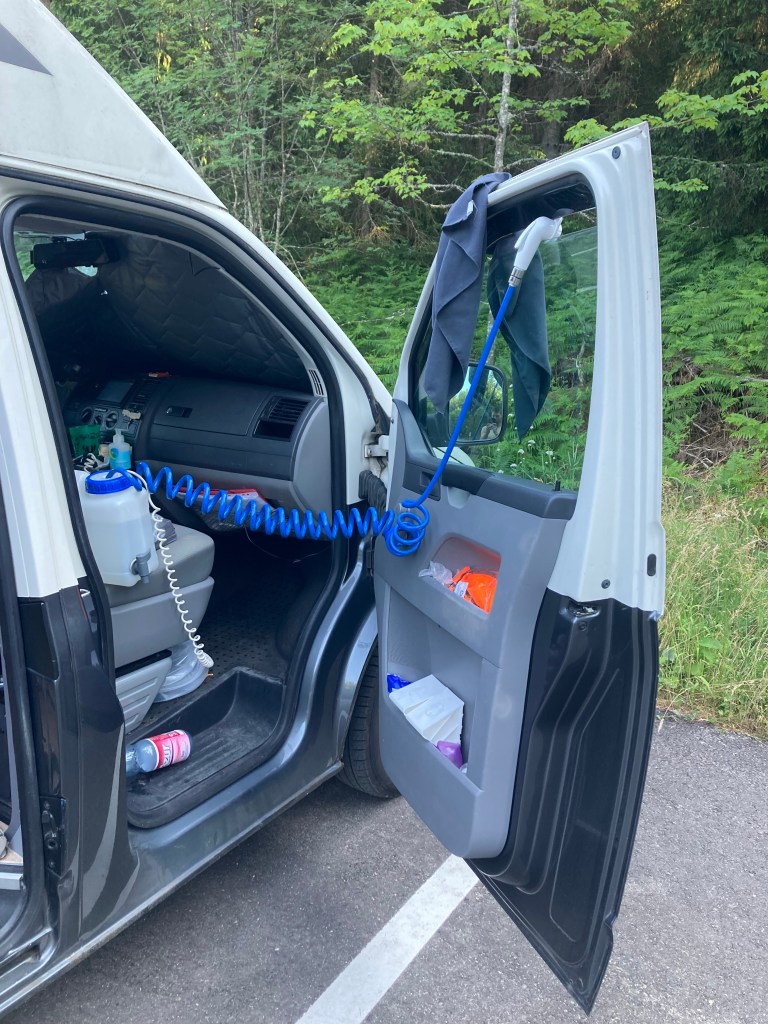 A picture of a van with the door open and a camping shower on the front seat with the camping shower hose poking through the window ready to be used